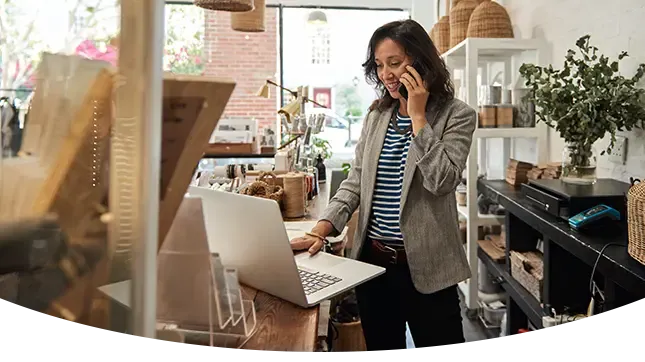 A woman business owner talks on a smartphone while using a laptop behind the counter of a boutique shop filled with home decor.